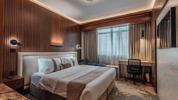 A hotel room with a large bed, a desk, and a chair. There is a window with curtains, a wall-mounted light, and a mirror.