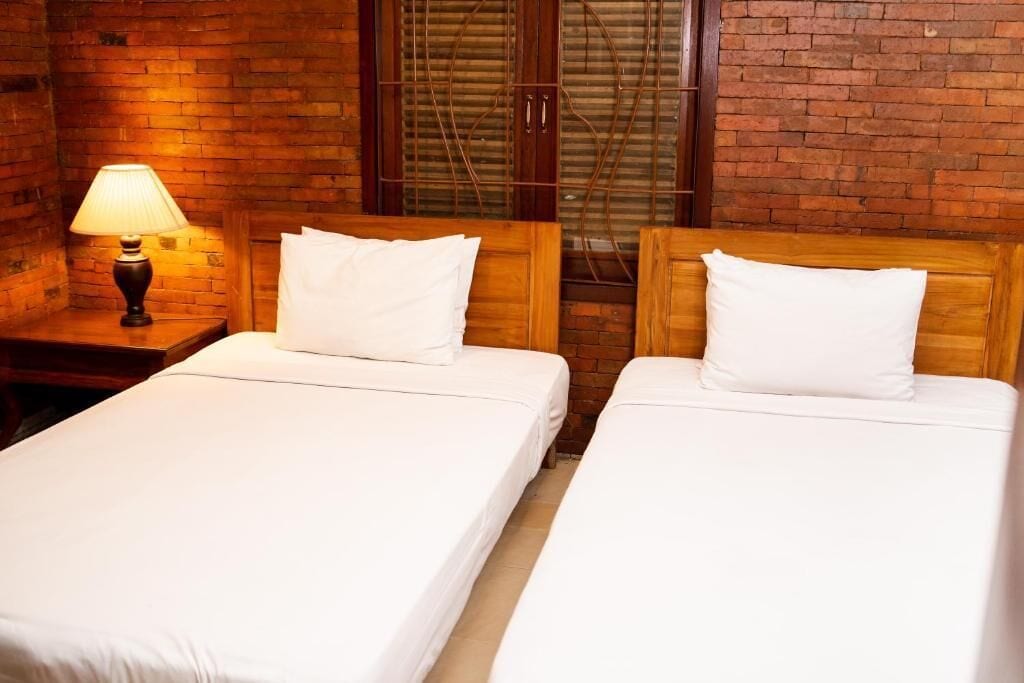 A room with two beds, a nightstand with a lamp, and a brick wall with a window.