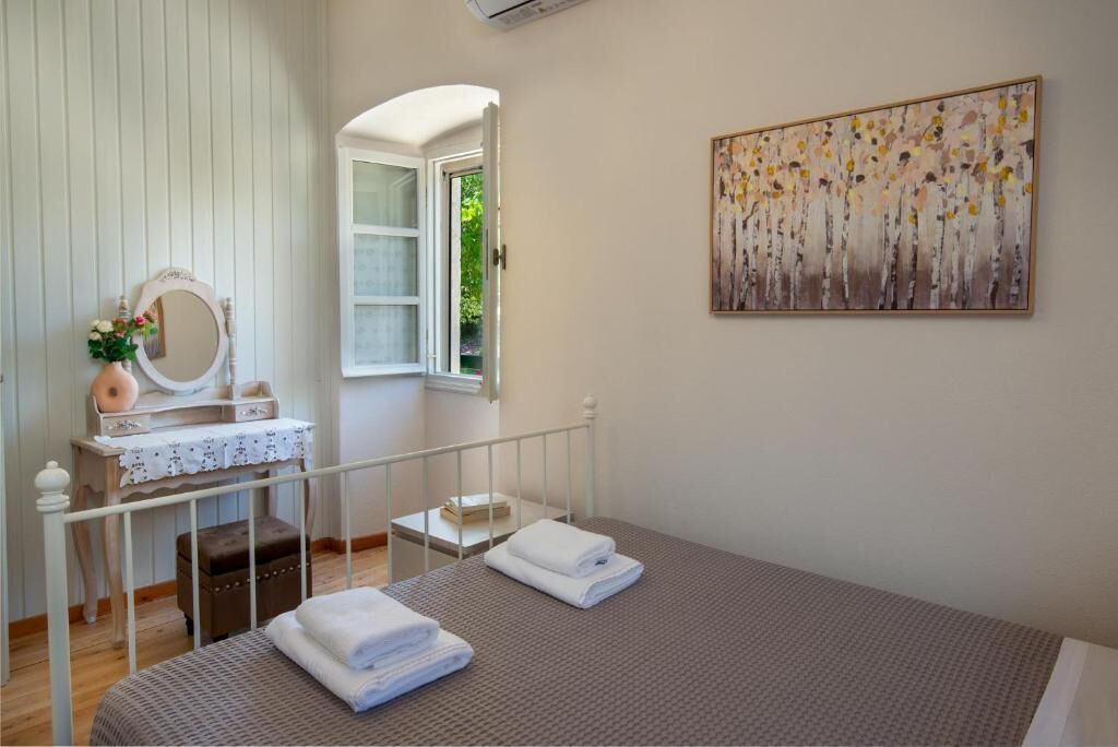 Un dormitorio con cama, una mesita de noche con espejo, una ventana y un cuadro en la pared.