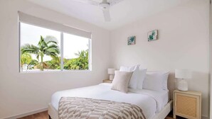 A bedroom with a bed, bedside lamps, a window with a view of palm trees, and a ceiling fan. - A Calm City Base Cairns CBD (Cairns North)