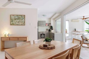 Un espace de salle à manger moderne avec une table en bois, des chaises, un ventilateur de plafond et une cuisine en arrière-plan.