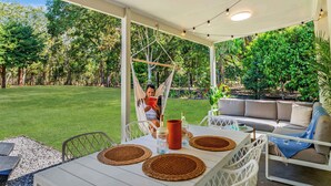 Un espacio de descanso al aire libre con techo, que incluye una hamaca, un sofá y una mesa con individuales y un florero.