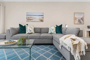 A living room with a gray sofa, a glass coffee table, a blue patterned rug, and two beach-themed wall art pieces.