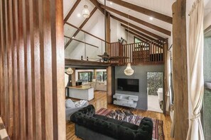 Uma sala de estar ampla com teto alto, vigas de madeira, um sofá preto, uma televisão e uma escada para um andar superior.
