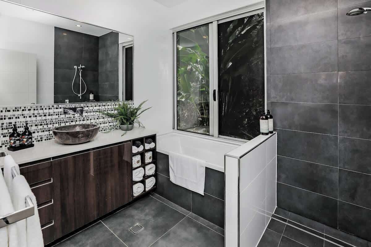A modern bathroom with a large mirror, a black and white tiled vanity, a large bowl sink, and a window with a view of greenery.