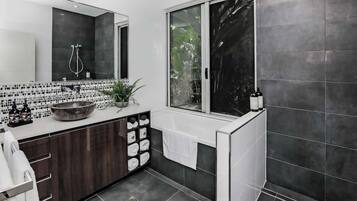 A modern bathroom with a large mirror, a black and white tiled vanity, a large bowl sink, and a window with a view of greenery.
