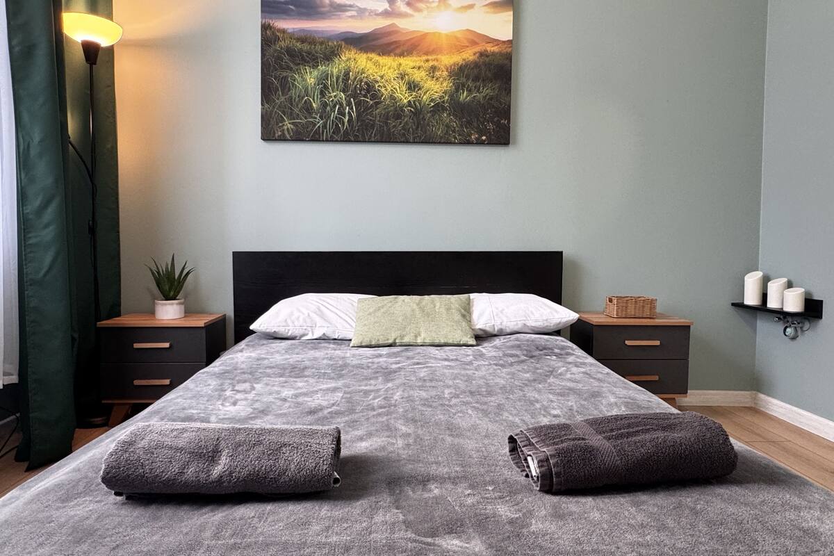 A bedroom with a bed, pillows, a nightstand with a lamp, a wall-mounted shelf, and a painting.