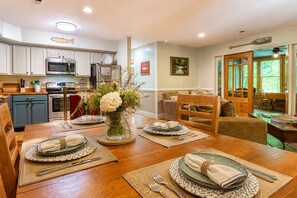 A dining area with a wooden table set for four, a vase of flowers, and a kitchen in the background. - Whispering Waters, Hot Tub, Riverside, Free Tickets (Pigeon Forge)