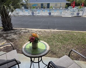 A patio with a table and chairs, a decorative vase with flowers, a palm tree, and a pool area in the background. - Designer #601 Chateau La Mer II 1st Floor 1-Bd 1 Bath Newly Added Near Pool (Destin)