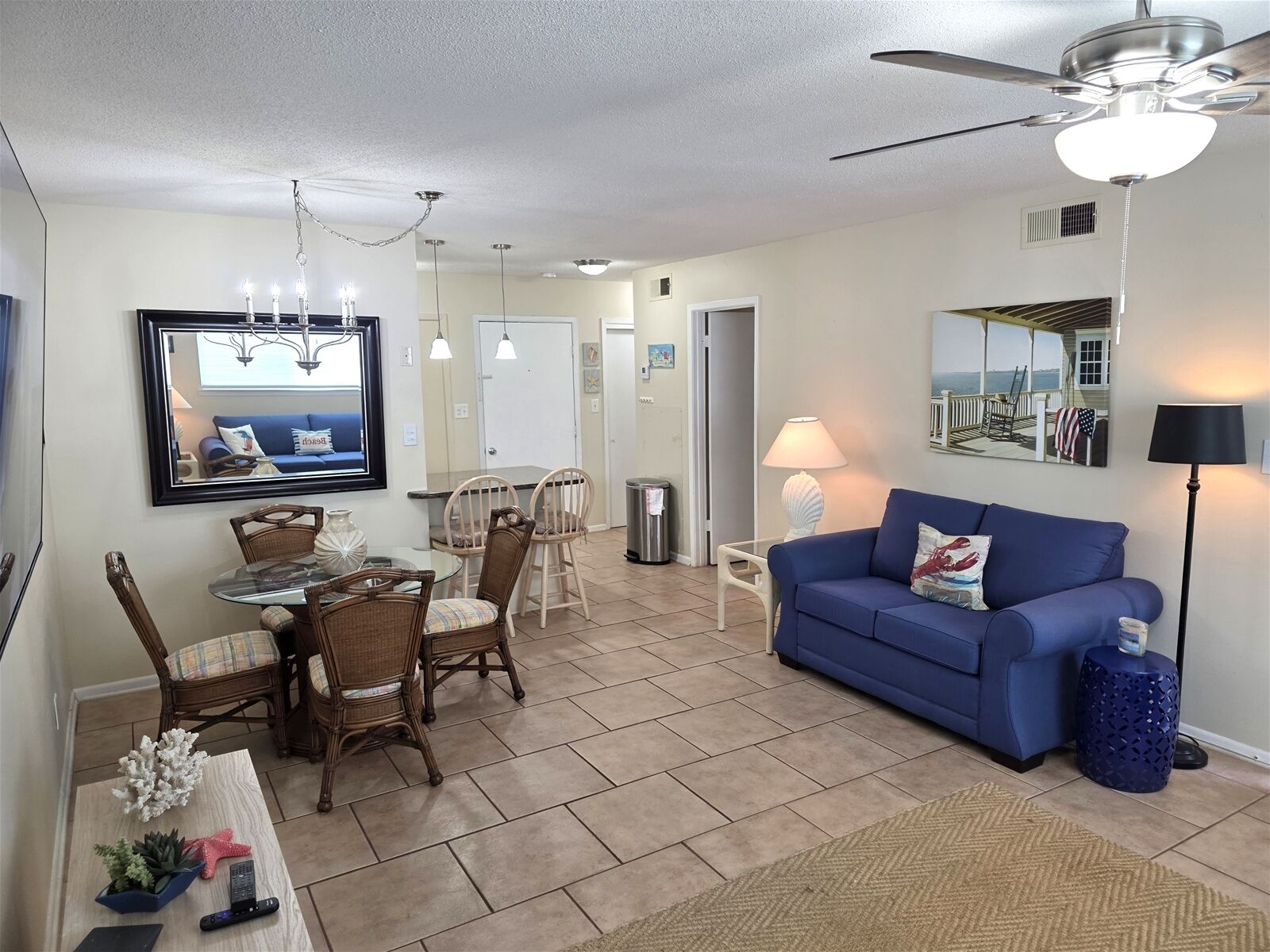 A living room with a blue sofa, a dining area with wicker chairs, a ceiling fan, and a large mirror on the wall.
