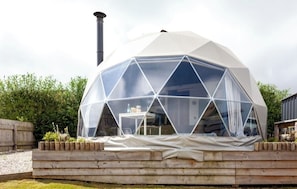 A geodesic dome structure with a chimney, surrounded by a wooden deck and greenery.