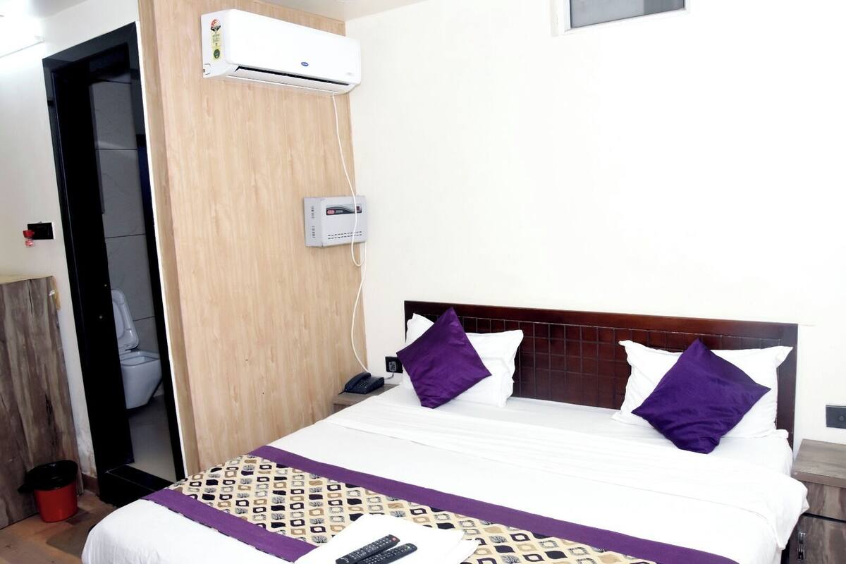 A hotel room with a bed, a bedside table, a phone, a remote control, an air conditioning unit, and a wall-mounted thermostat.