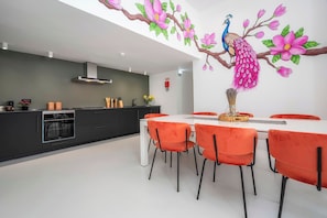 A modern kitchen with a dining area, featuring a peacock mural, a white dining table, and orange chairs. - Marina Legacy I Demi-suite Infinity 8 (Faro)