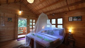A wooden cabin bedroom with a bed under a mosquito net, wooden walls, and a large window.