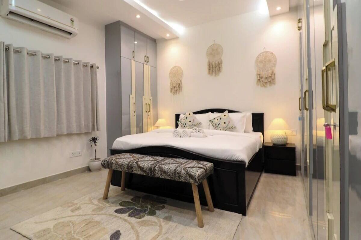 A bedroom with a bed, bedside tables, a bench, a wall-mounted air conditioner, and a window with curtains.