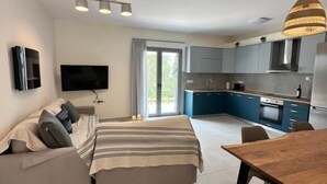 A modern living room with a sofa, a TV, a dining table with chairs, and a kitchen area with cabinets and a stove. - Scandal House by All About Paxos (Paxos)