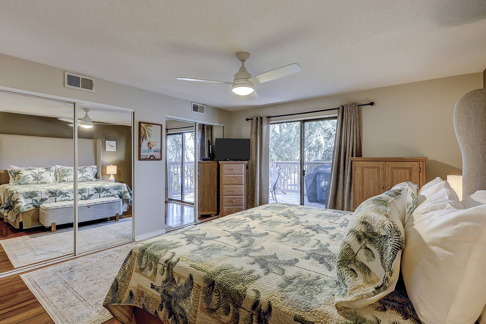 A bedroom with a bed, a dresser, a television, a ceiling fan, and a sliding glass door leading to a balcony.