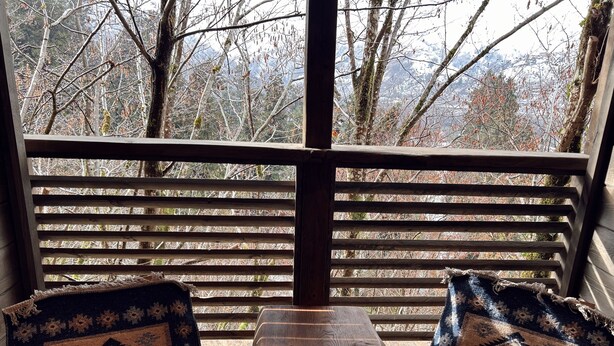 A wooden bench with a small table, two patterned chairs, and a wooden railing with a view of trees and mountains.