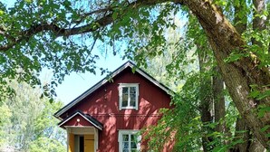 Une maison en bois rouge, avec une porte jaune et des boiseries blanches, entourée de verdure et d’arbres.