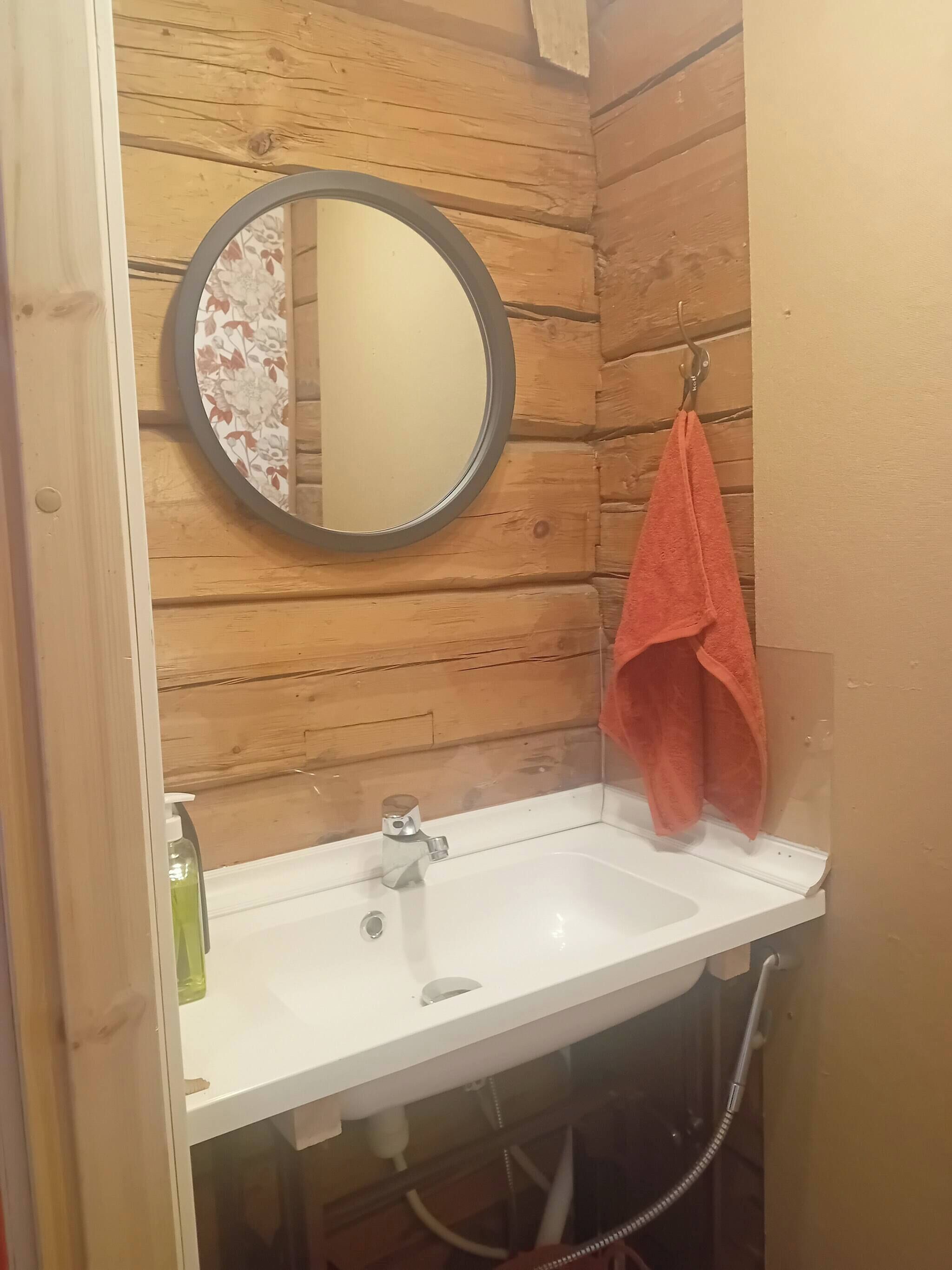 A bathroom with a wooden wall, a round mirror, a white sink, and a red towel hanging on a hook.