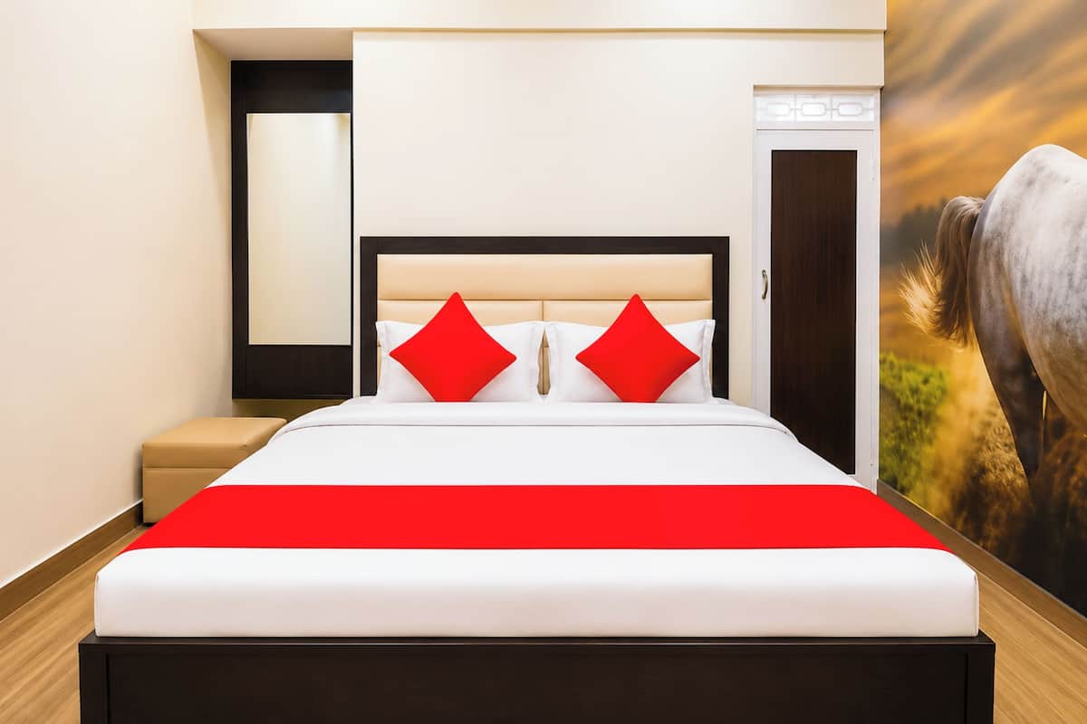 A hotel room with a bed, a red accent pillow, a beige ottoman, and a door.