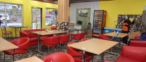 A restaurant interior with red chairs, wooden tables, a Coca-Cola refrigerator, and a person standing near the counter.