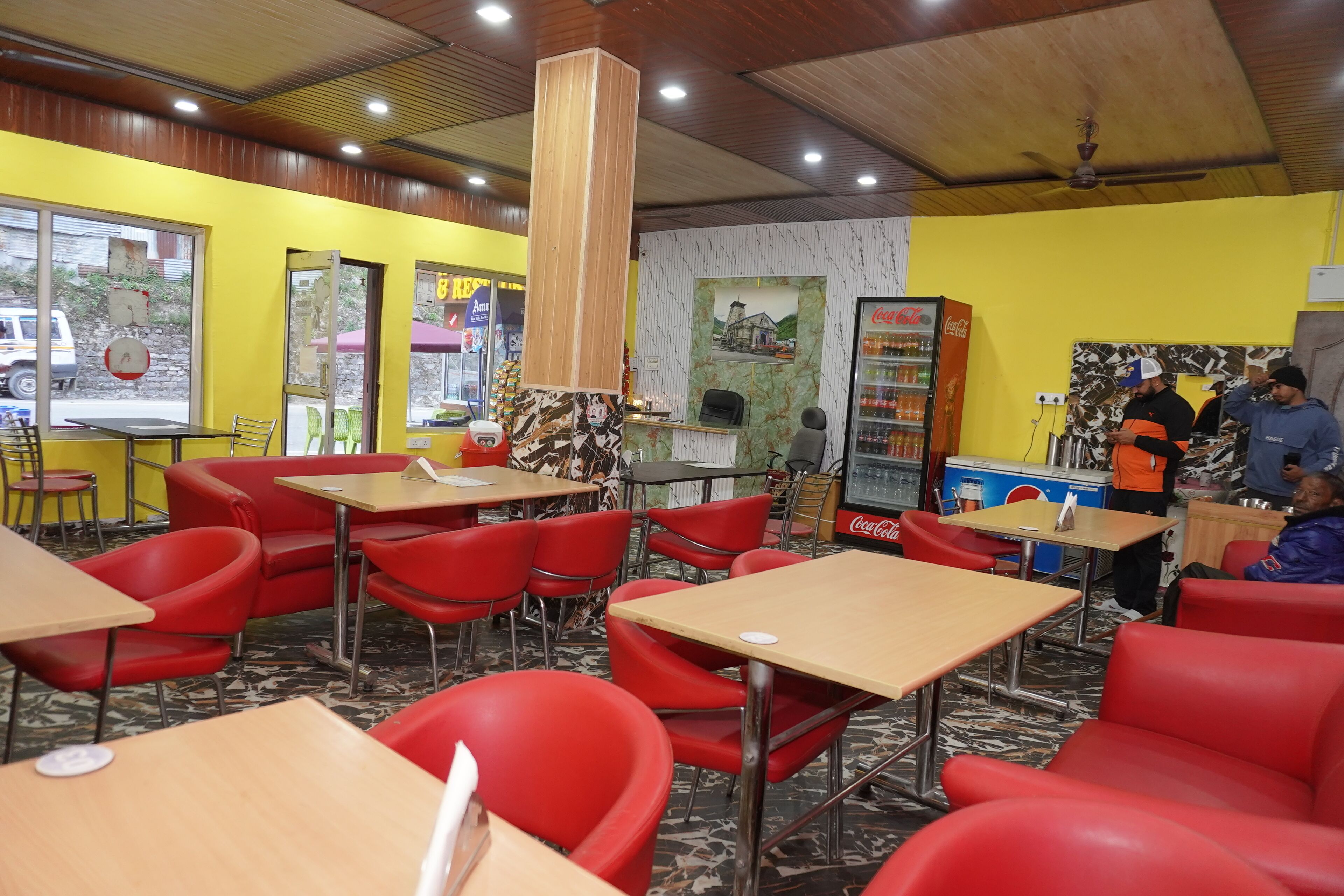 A restaurant with red chairs and tables, a Coca-Cola refrigerator, and a wooden pillar.