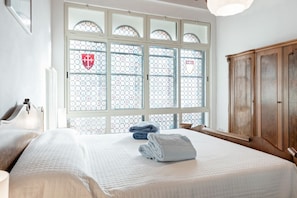 Una camera d'albergo con una grande finestra, un letto con copripiumino bianco, un armadio in legno e una testiera.