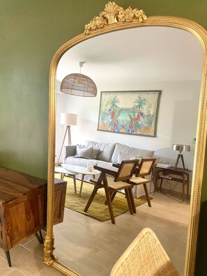 Ein Wohnzimmer mit einem großen, verzierten Spiegel, einem Sofa, einem Couchtisch, einem Korbstuhl, einer Stehlampe und einem Bild an der Wand.