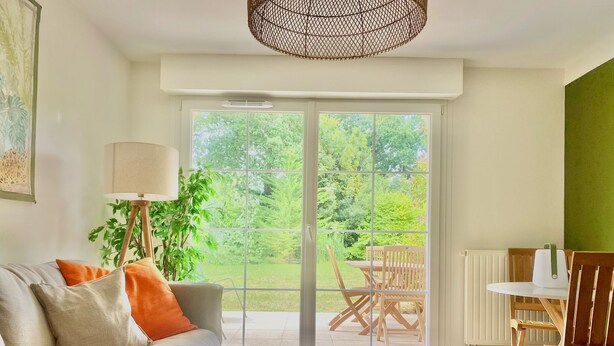 Ein Wohnzimmer mit einer Couch, einem Couchtisch, einer Lampe, einem Esstisch mit Stühlen und Blick durch eine große Glasschiebetür auf einen Garten.