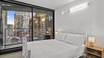 Habitación de hotel moderna con una cama grande, una mesita de noche, una lámpara y vistas a un paisaje urbano a través de ventanas del suelo al techo.