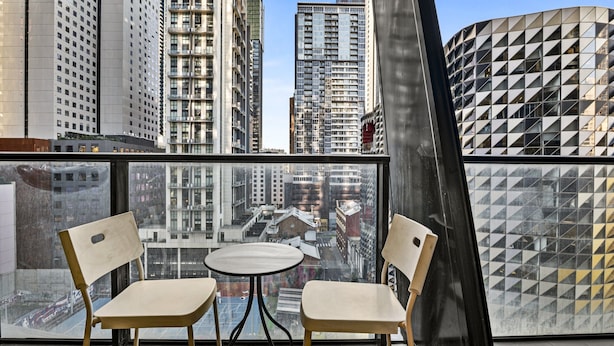A balcony with two chairs and a small table, overlooking a cityscape with tall buildings and a distinctive geometric building. - Central 2 Bed Apartment Near Melbourne CBD (Melbourne)