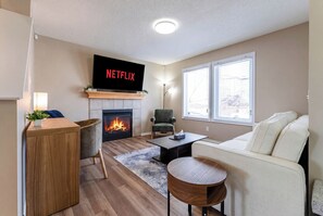 Una sala de estar con chimenea, televisor montado encima, sofá, mesa de centro y una silla.
