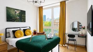 Un dormitorio con cama, escritorio, silla, un cuadro y vistas a edificios.