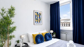 Una habitación de hotel moderna con una cama, una mesita de noche con una lámpara, una planta en maceta, una ventana con cortinas y vistas a un edificio.