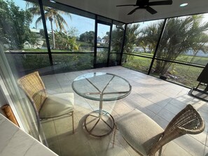 A glass-enclosed balcony with a round glass table and wicker chairs, overlooking a garden with palm trees.