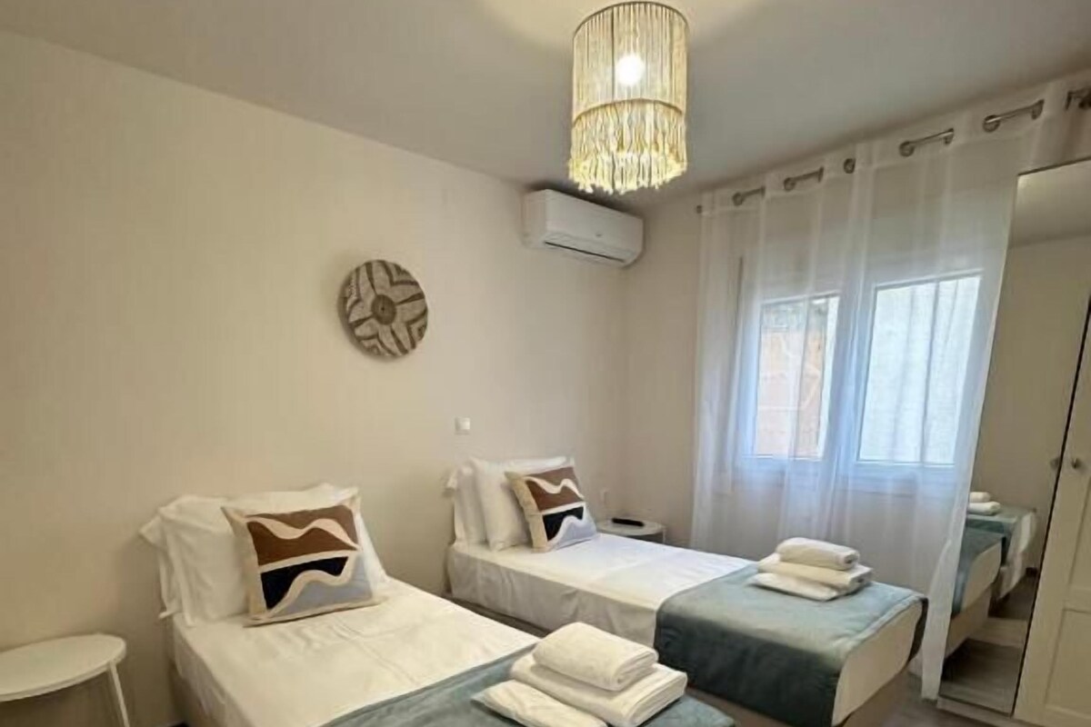 A hotel room with two single beds, a small table, a chandelier, an air conditioning unit, and a view of the outside.
