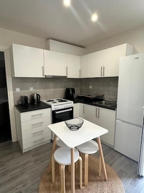 A compact kitchen with white cabinets, a stove, a coffee maker, and a small table with chairs. - Rouli's Houses by All About Paxos (Paxos)