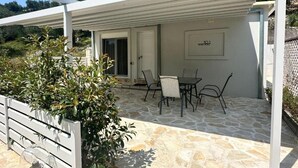 A covered outdoor patio with stone flooring, a table with chairs, and a door leading inside. - Rouli's Houses by All About Paxos (Paxos)