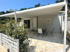 A covered outdoor patio with stone flooring, a table with chairs, and a door leading inside. - Rouli's Houses by All About Paxos (Paxos)