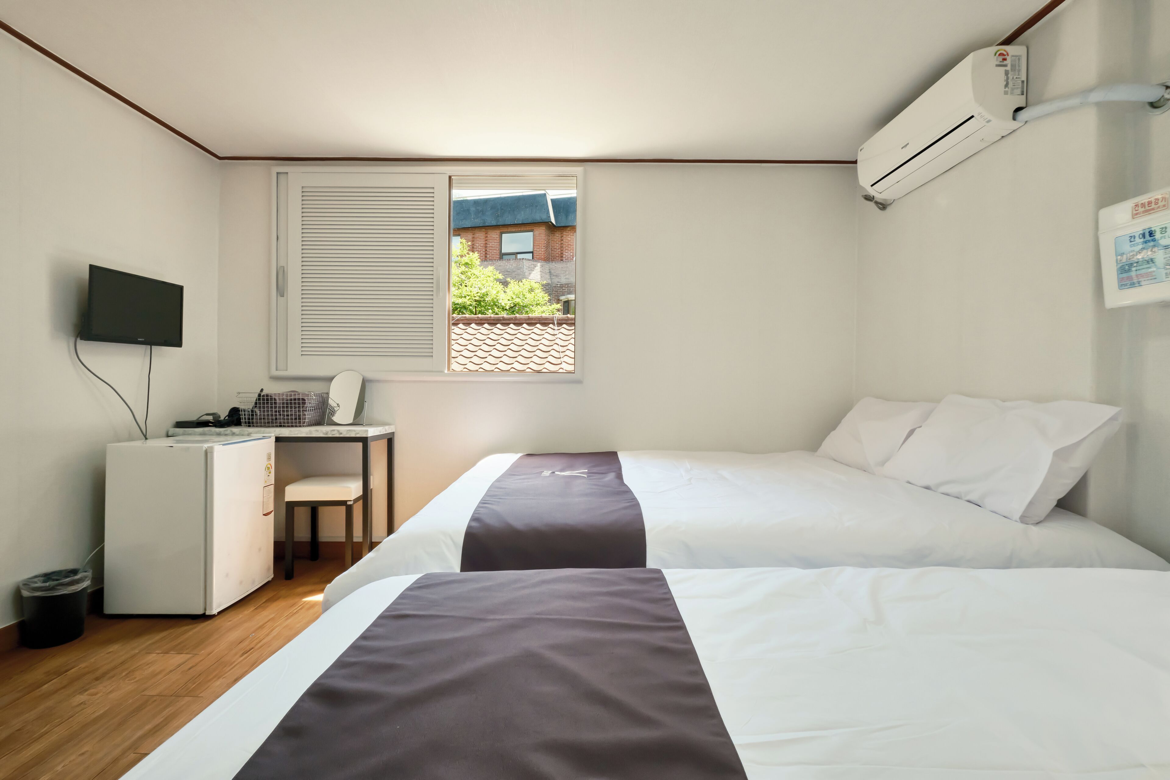 A hotel room with two beds, a desk, a chair, a TV, an air conditioner, and a window with blinds.