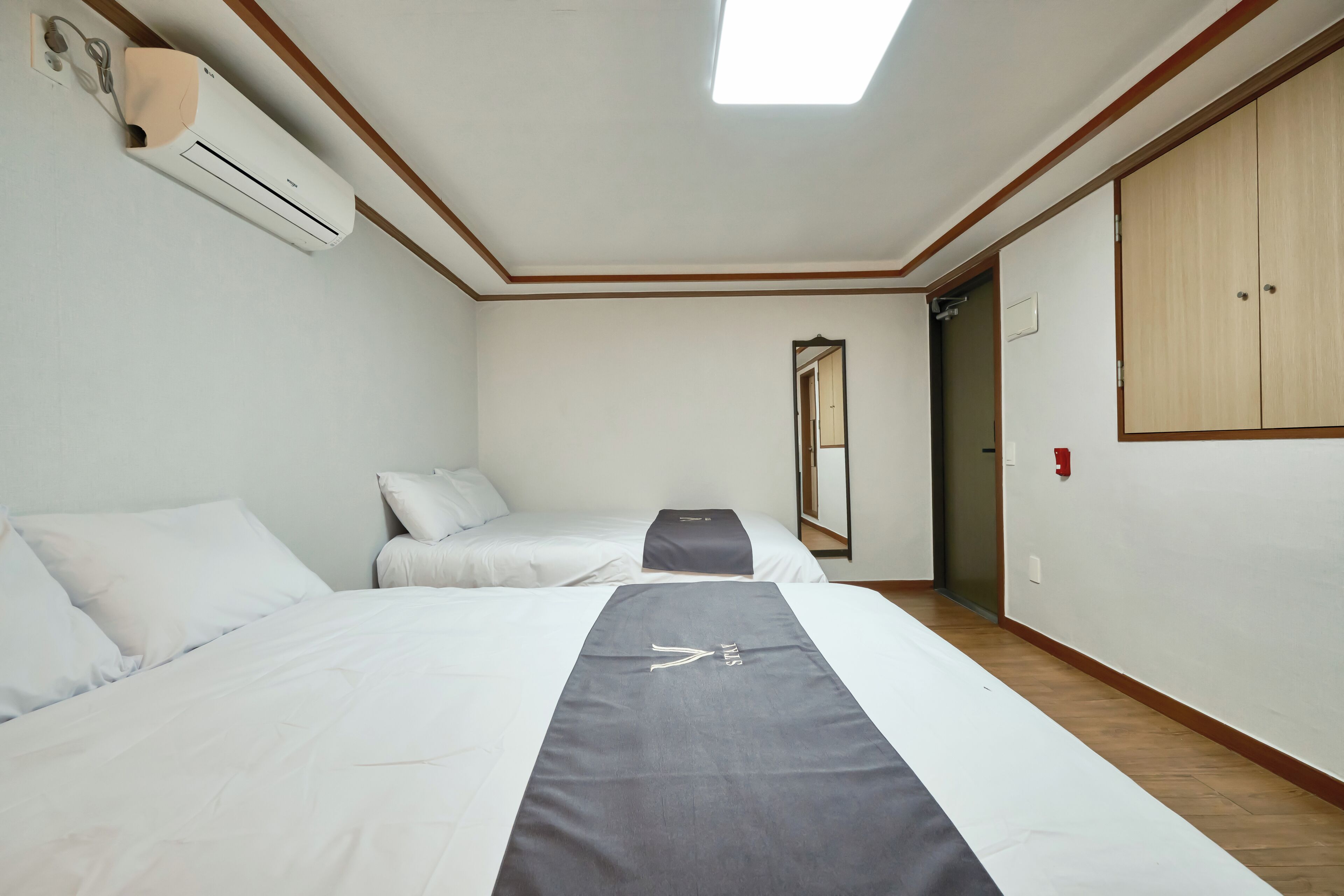 A hotel room with two beds, a ceiling fan, a wall-mounted air conditioner, a mirror, and a wardrobe.