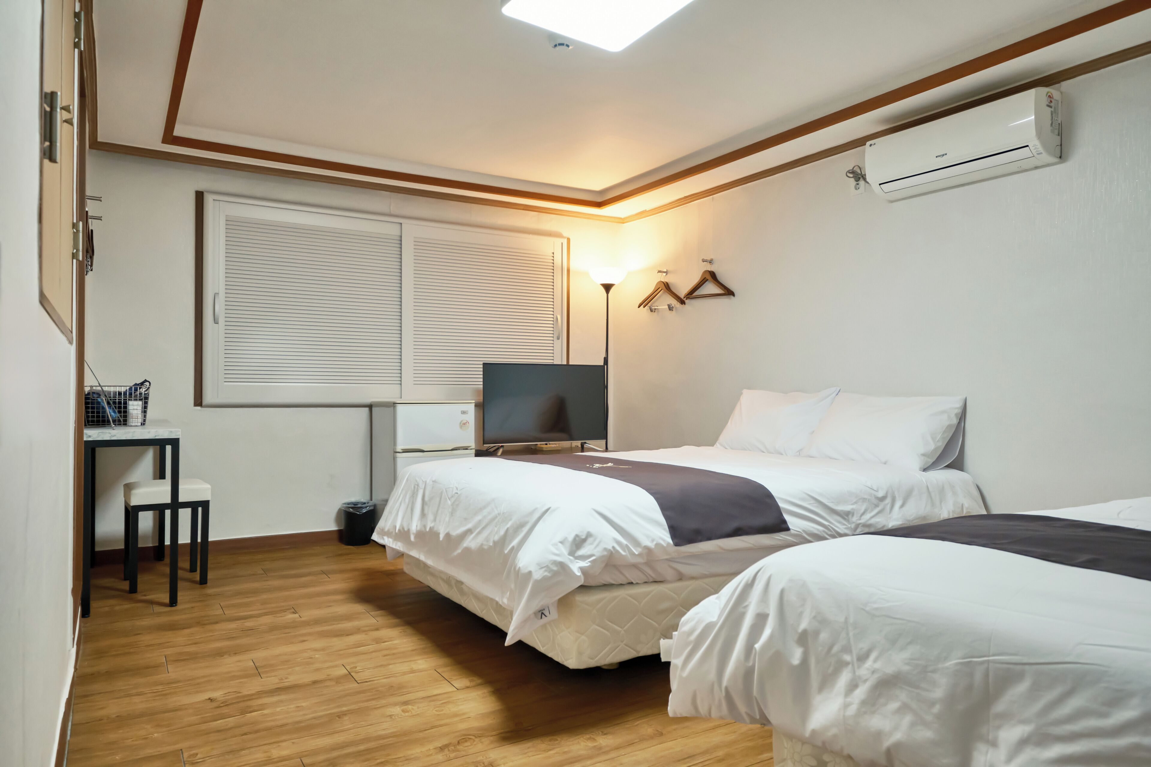 A hotel room with two beds, a desk, a TV, and an air conditioning unit.