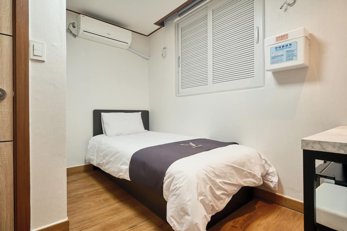 A small, single-bed room with a wooden floor, a window with blinds, an air conditioning unit, and a bedside table.