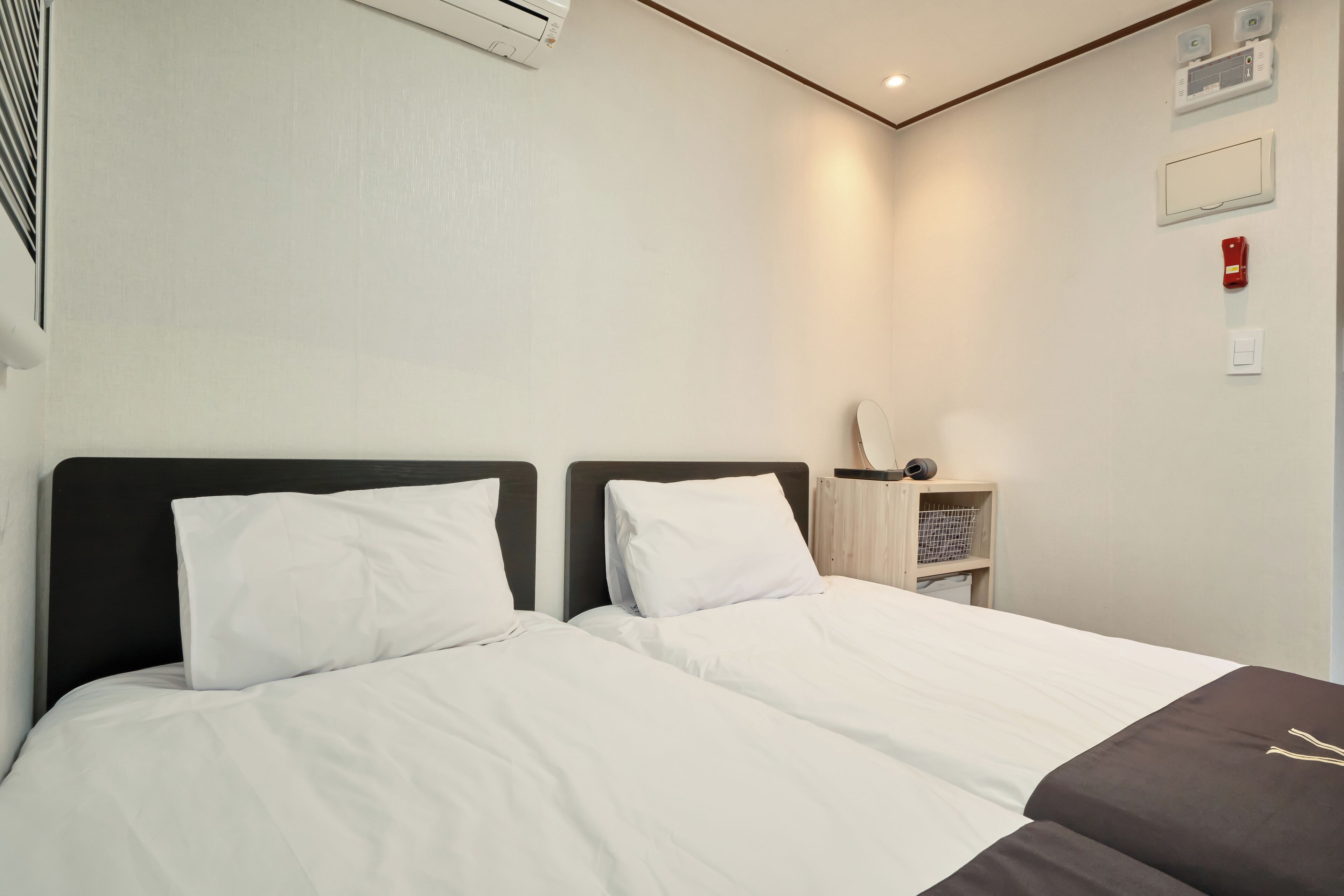 Une chambre d’hôtel avec deux lits, une petite table de chevet en bois, un climatiseur fixé au mur et un détecteur d’incendie.