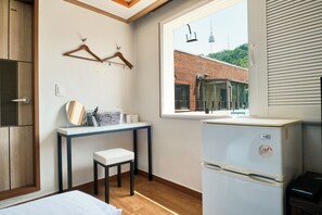 Habitación compacta con cama, una mesita con silla, refrigerador blanco y ventana con vista a un edificio y una torre.