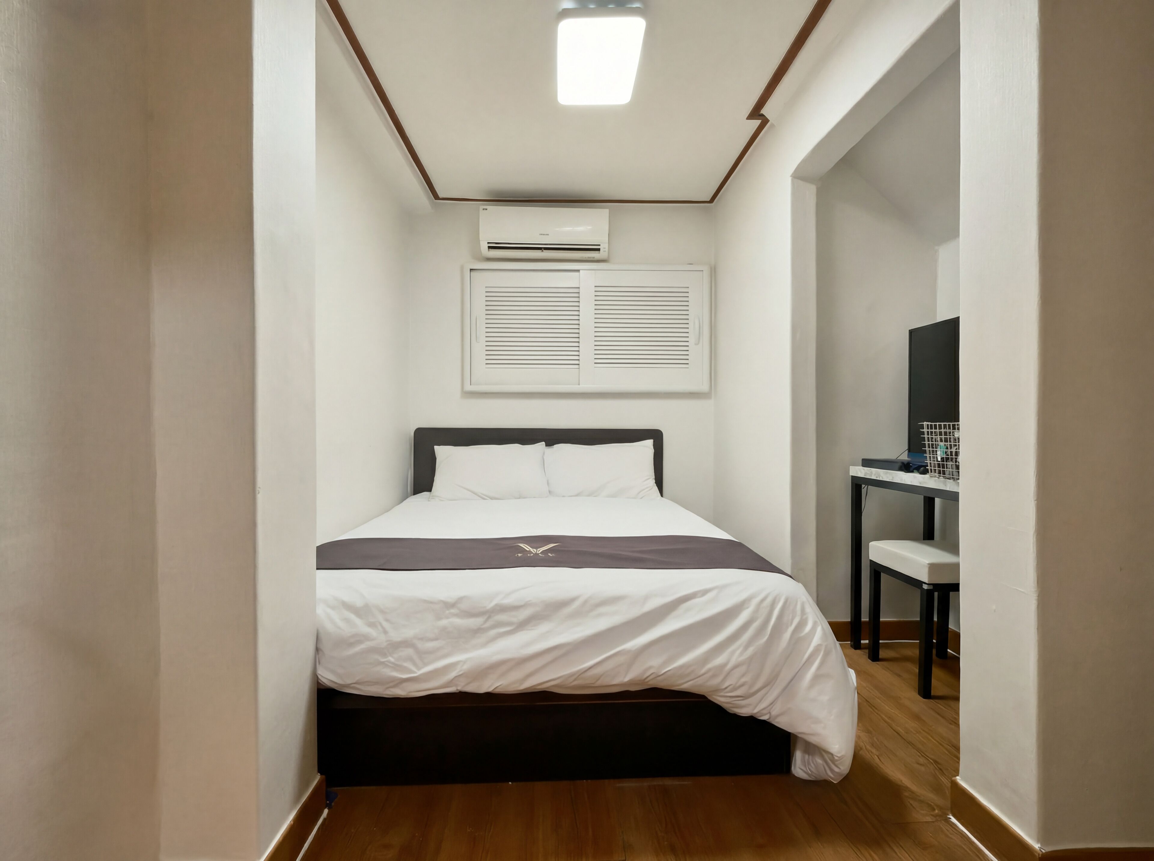 A narrow room with a bed, a desk, and an air conditioning unit.