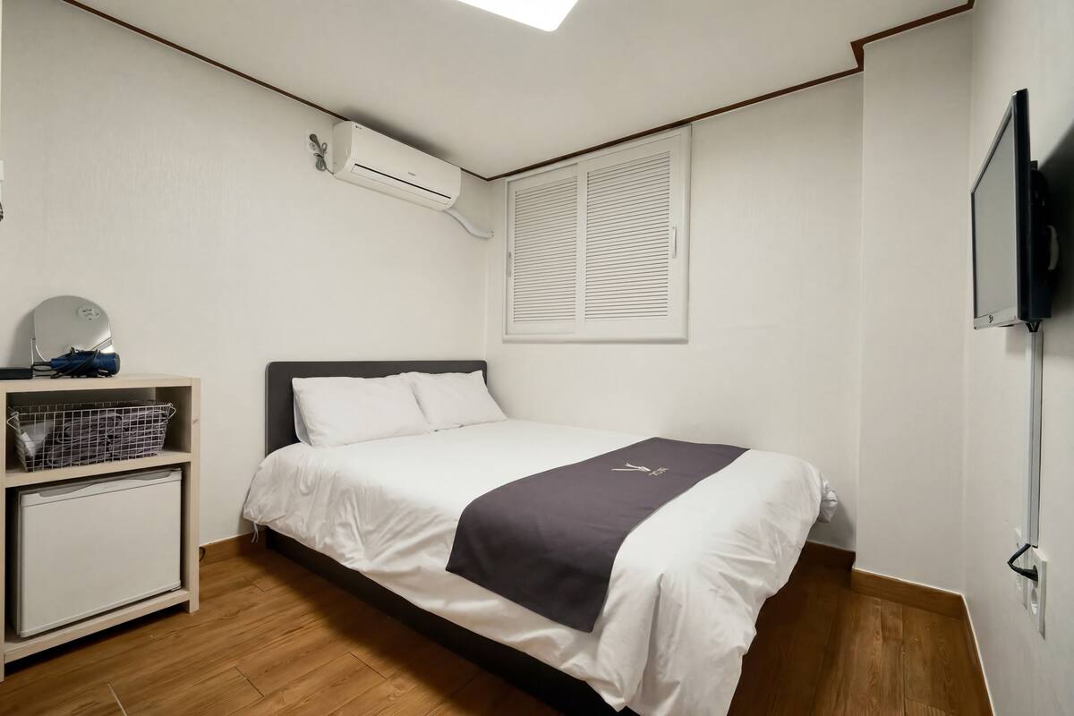 A bedroom with a bed, a shelf with a basket, a wall-mounted air conditioner, and a window with blinds.