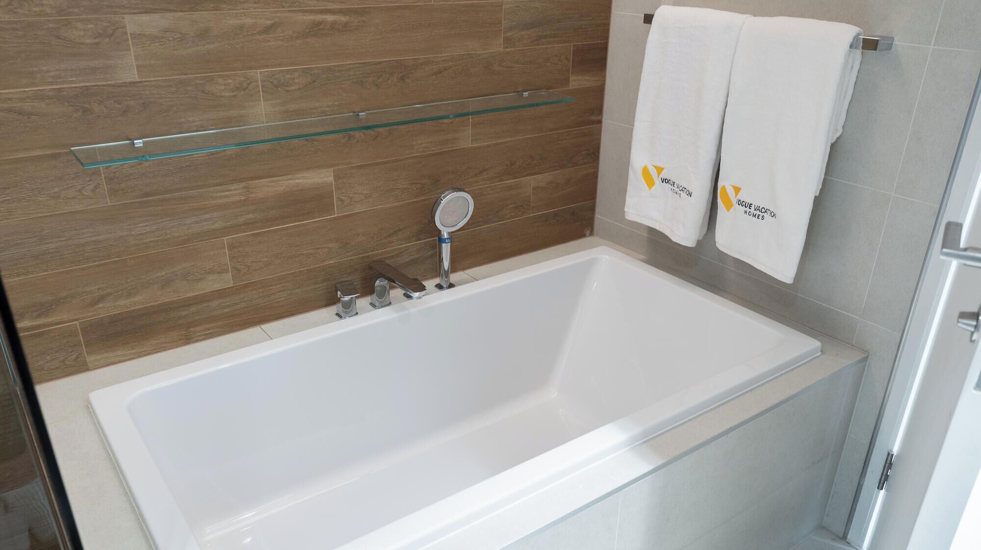 Un baño moderno con una bañera blanca, revestimiento de madera en las paredes, un estante de vidrio y dos toallas con un logotipo.