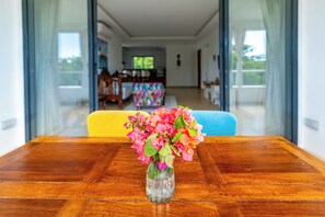 Ein Holztisch mit einer klaren Glasvase mit rosafarbenen Blumen, ein blaues Sofa und ein farbig gemustertes Sofa in einem hellen Raum mit großen Fenstern.
